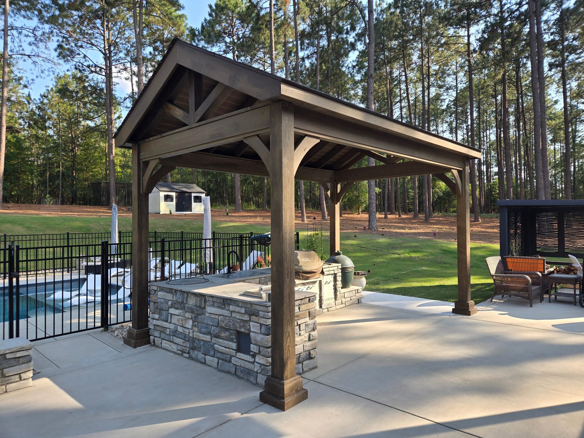 Wood pavilion and stone outdoor kitchen beside a pool, backed by tall Carolina pines at golden hour